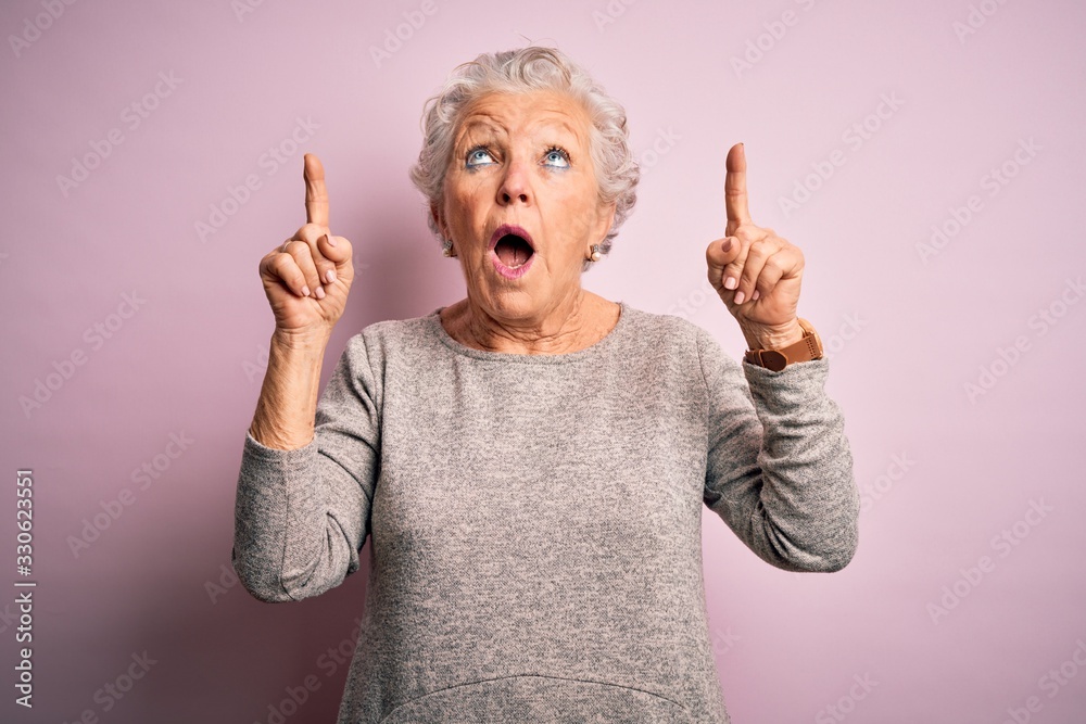 Senior beautiful woman wearing casual t-shirt standing over isolated pink background amazed and surprised looking up and pointing with fingers and raised arms.