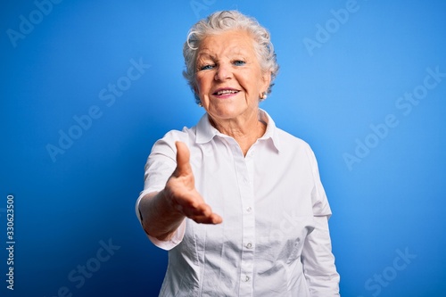 Senior beautiful woman wearing elegant shirt standing over isolated blue background smiling friendly offering handshake as greeting and welcoming. Successful business.