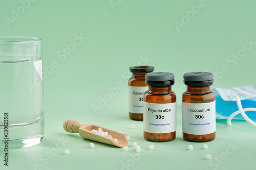 Bottles of homeopathic drugs - Bryonia, Lycopodium, Gelsemium - recommended by homeopaths for treatment and prophylaxis of Coronavirus Covid-19, glass of water, scoop, medical protective mask. photo