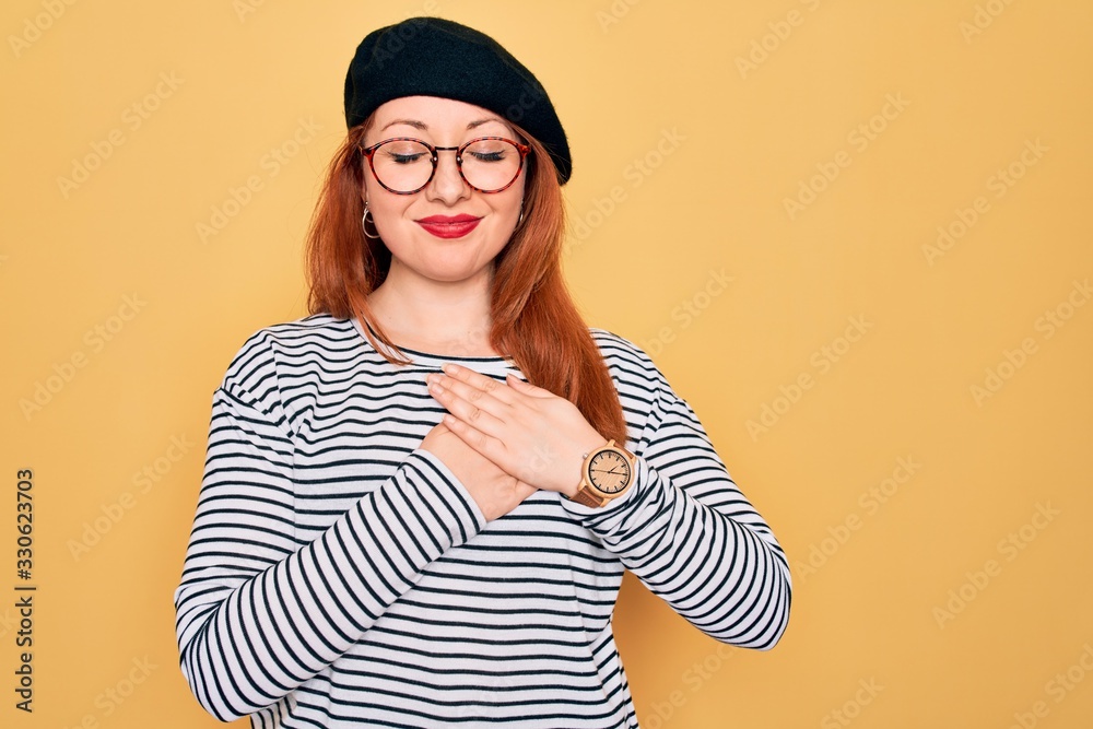 Beautiful redhead woman wearing striped t-shirt and french beret over yellow background smiling with hands on chest with closed eyes and grateful gesture on face. Health concept.