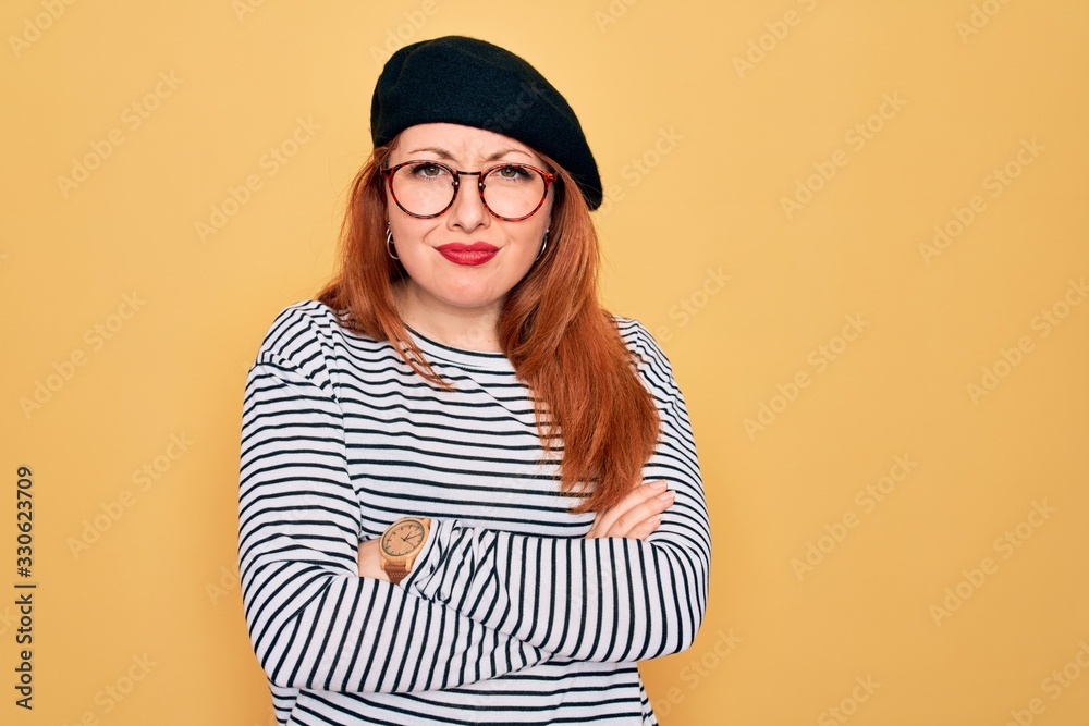 Beautiful redhead woman wearing striped t-shirt and french beret over yellow background skeptic and nervous, disapproving expression on face with crossed arms. Negative person.