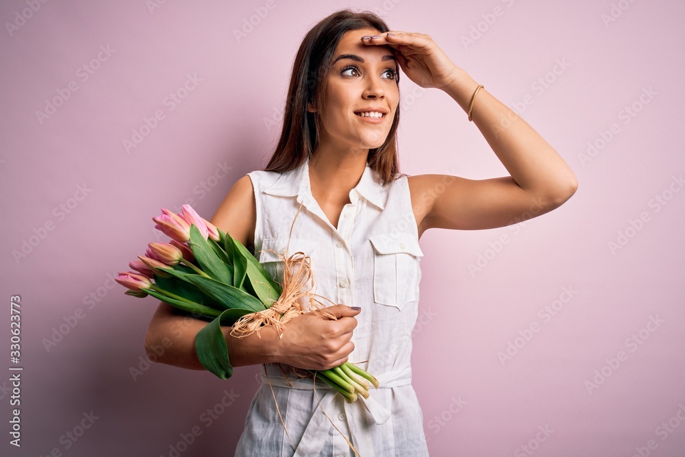 Young beautiful brunette woman holding bouquet of tulips flowers over pink background very happy and smiling looking far away with hand over head. Searching concept. Young beautiful brunette woman holding bouquet of tulips flowers over pink background very happy and smiling looking far away with hand over head. Searching concept.
