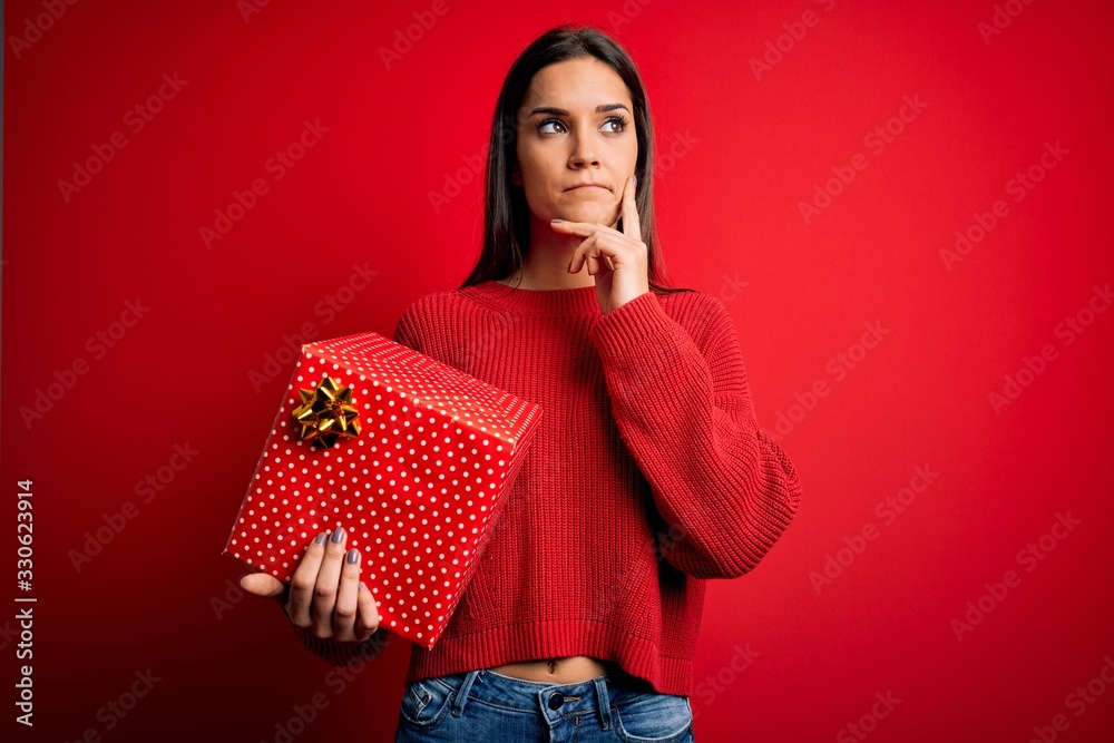 Young beautiful brunette woman holding birthday gift over isolated red background serious face thinking about question, very confused idea Young beautiful brunette woman holding birthday gift over isolated red background serious face thinking about question, very confused idea