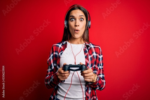 Young beautiful brunette gamer woman playing video game using joystick and headphones scared in shock with a surprise face, afraid and excited with fear expression photo
