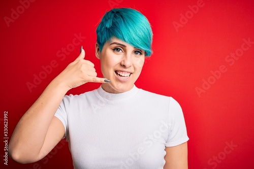 Young beautiful woman with blue fashion hair wearing casual t-shirt over red background smiling doing phone gesture with hand and fingers like talking on the telephone. Communicating concepts.