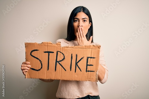 Young beautiful hispanic woman holding protest banner at strike or manifestation cover mouth with hand shocked with shame for mistake, expression of fear, scared in silence, secret concept © Krakenimages.com
