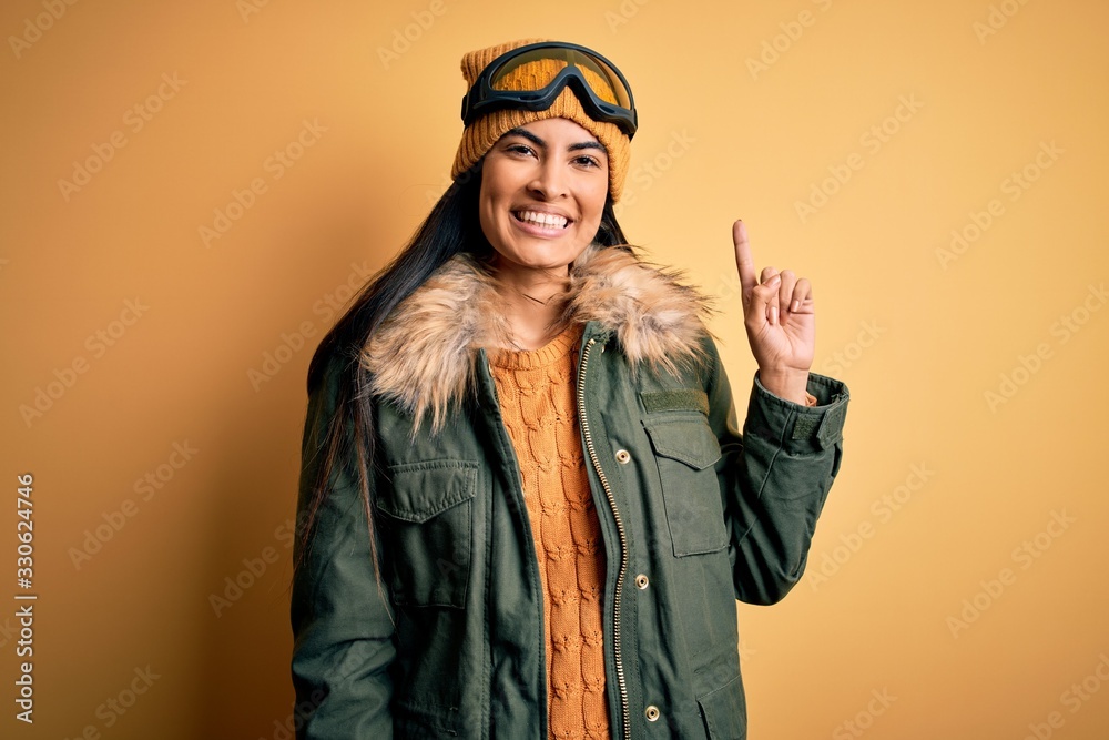 Young beautiful hispanic woman wearing ski glasses and coat for winter weather showing and pointing up with finger number one while smiling confident and happy.