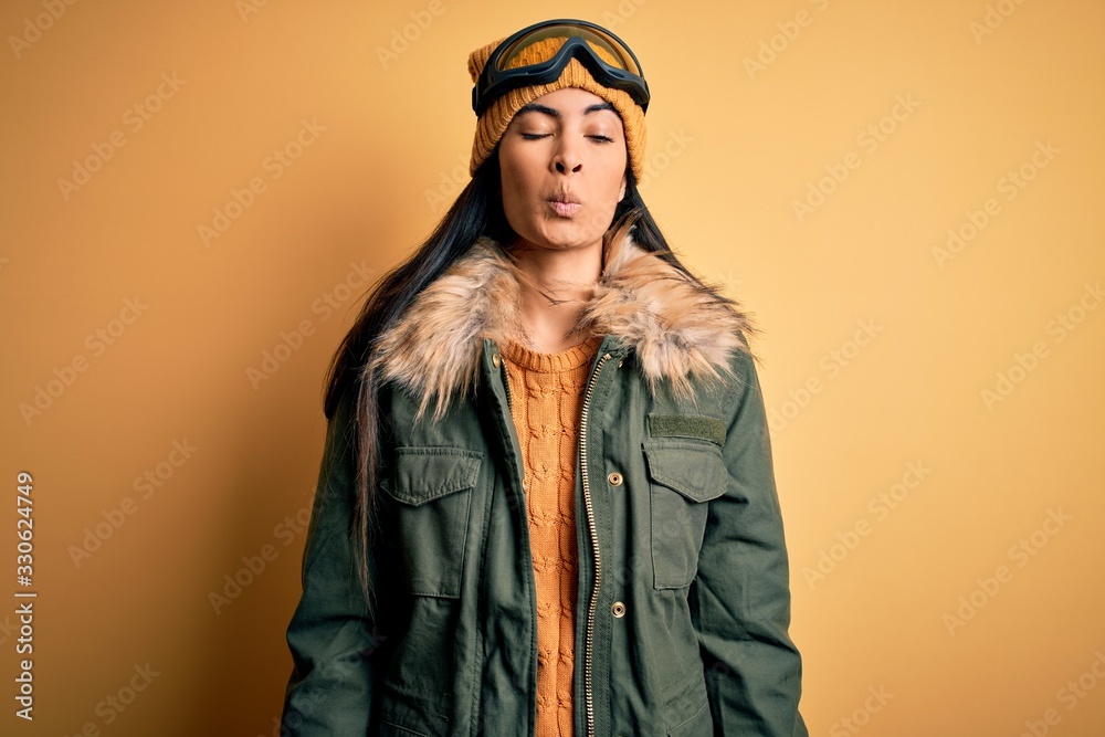 Young beautiful hispanic woman wearing ski glasses and coat for winter weather making fish face with lips, crazy and comical gesture. Funny expression.