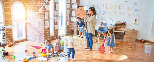 Obraz Beautiful teacher and group of toddlers playing around lots of toys at kindergarten
