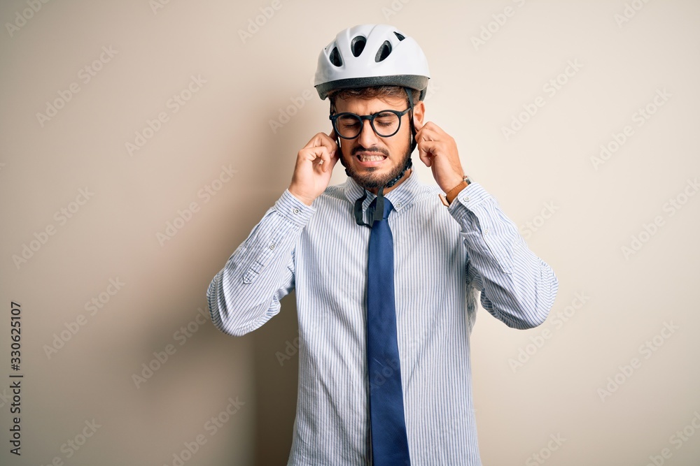 Young businessman wearing glasses and bike helmet standing over isolated white bakground covering ears with fingers with annoyed expression for the noise of loud music. Deaf concept. Young businessman wearing glasses and bike helmet standing over isolated white bakground covering ears with fingers with annoyed expression for the noise of loud music. Deaf concept.