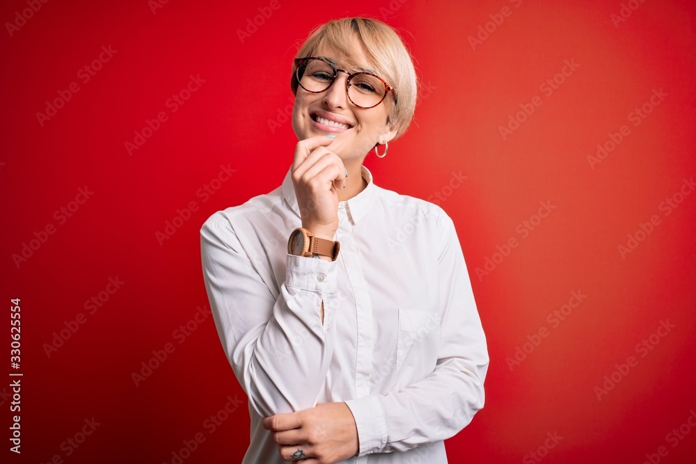 Young blonde business woman with short hair wearing glasses over red background looking confident at the camera with smile with crossed arms and hand raised on chin. Thinking positive. Young blonde business woman with short hair wearing glasses over red background looking confident at the camera with smile with crossed arms and hand raised on chin. Thinking positive.