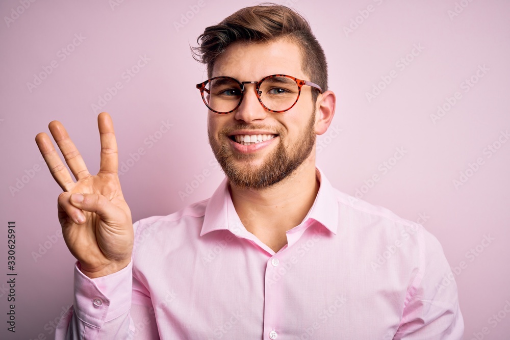 Young handsome blond man with beard and blue eyes wearing pink shirt and glasses showing and pointing up with fingers number three while smiling confident and happy. Young handsome blond man with beard and blue eyes wearing pink shirt and glasses showing and pointing up with fingers number three while smiling confident and happy.
