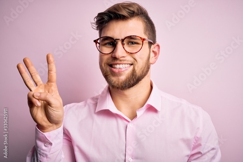 Young handsome blond man with beard and blue eyes wearing pink shirt and glasses showing and pointing up with fingers number three while smiling confident and happy.