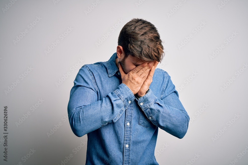 Young handsome blond man with beard and blue eyes wearing casual denim shirt with sad expression covering face with hands while crying. Depression concept. Young handsome blond man with beard and blue eyes wearing casual denim shirt with sad expression covering face with hands while crying. Depression concept.