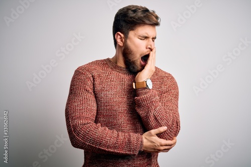 Young blond man with beard and blue eyes wearing casual sweater over white background bored yawning tired covering mouth with hand. Restless and sleepiness.