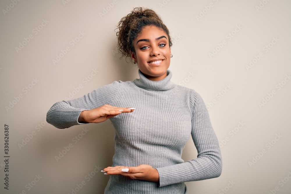 Beautiful african american girl wearing turtleneck sweater standing over white background gesturing with hands showing big and large size sign, measure symbol. Smiling looking at the camera. Measuring Beautiful african american girl wearing turtleneck sweater standing over white background gesturing with hands showing big and large size sign, measure symbol. Smiling looking at the camera. Measuring