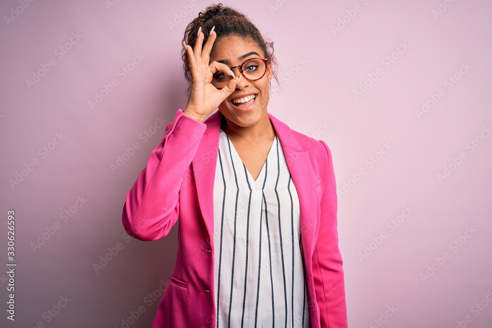 Beautiful african american businesswoman wearing jacket and glasses over pink background doing ok gesture with hand smiling, eye looking through fingers with happy face.