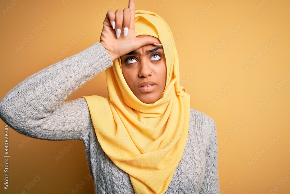 Young beautiful african american girl wearing muslim hijab over isolated yellow background making fun of people with fingers on forehead doing loser gesture mocking and insulting.