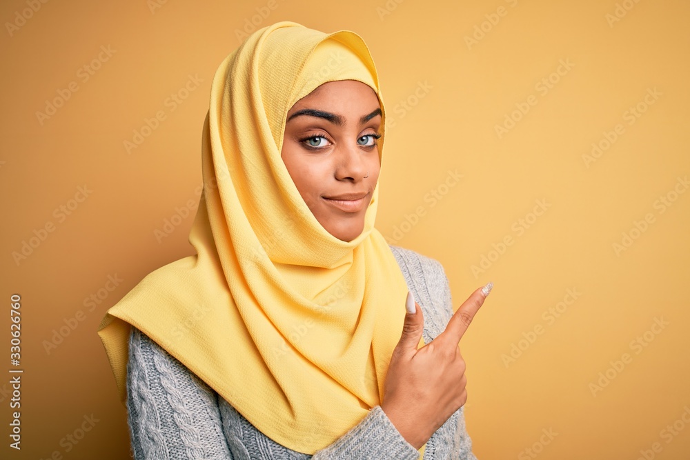 Young beautiful african american girl wearing muslim hijab over isolated yellow background Pointing with hand finger to the side showing advertisement, serious and calm face Young beautiful african american girl wearing muslim hijab over isolated yellow background Pointing with hand finger to the side showing advertisement, serious and calm face