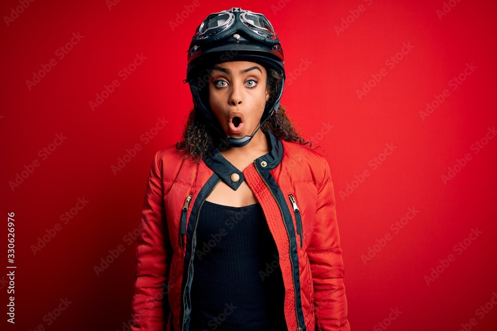 Young african american motorcyclist girl wearing moto helmet and glasses over red background afraid and shocked with surprise expression, fear and excited face. Young african american motorcyclist girl wearing moto helmet and glasses over red background afraid and shocked with surprise expression, fear and excited face.
