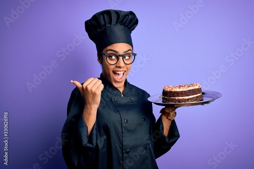 Young african american cooker girl wearing uniform and hat holding tray with cake pointing and showing with thumb up to the side with happy face smiling