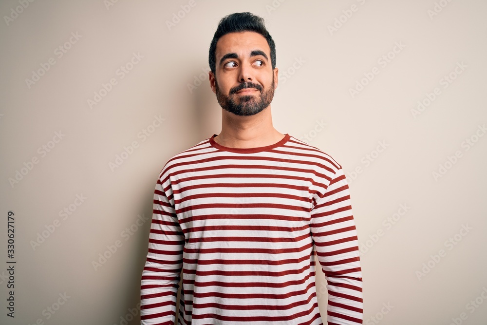 Young handsome man with beard wearing casual striped t-shirt standing over white background smiling looking to the side and staring away thinking.