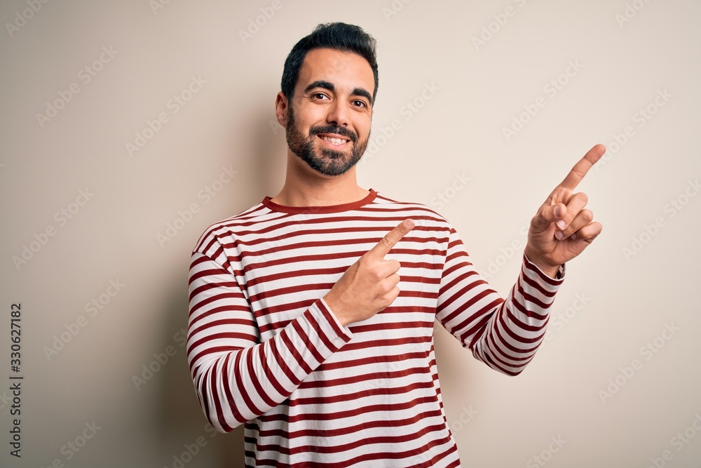 Young handsome man with beard wearing casual striped t-shirt standing over white background smiling and looking at the camera pointing with two hands and fingers to the side. Young handsome man with beard wearing casual striped t-shirt standing over white background smiling and looking at the camera pointing with two hands and fingers to the side.