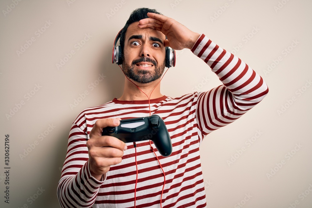 Young handsome gamer man with beard playing video game using joystick and headphones stressed with hand on head, shocked with shame and surprise face, angry and frustrated. Fear and upset for mistake.