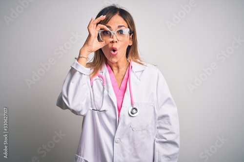 Middle age beautiful doctor woman wearing pink stethoscope over isolated white background doing ok gesture shocked with surprised face, eye looking through fingers. Unbelieving expression. photo