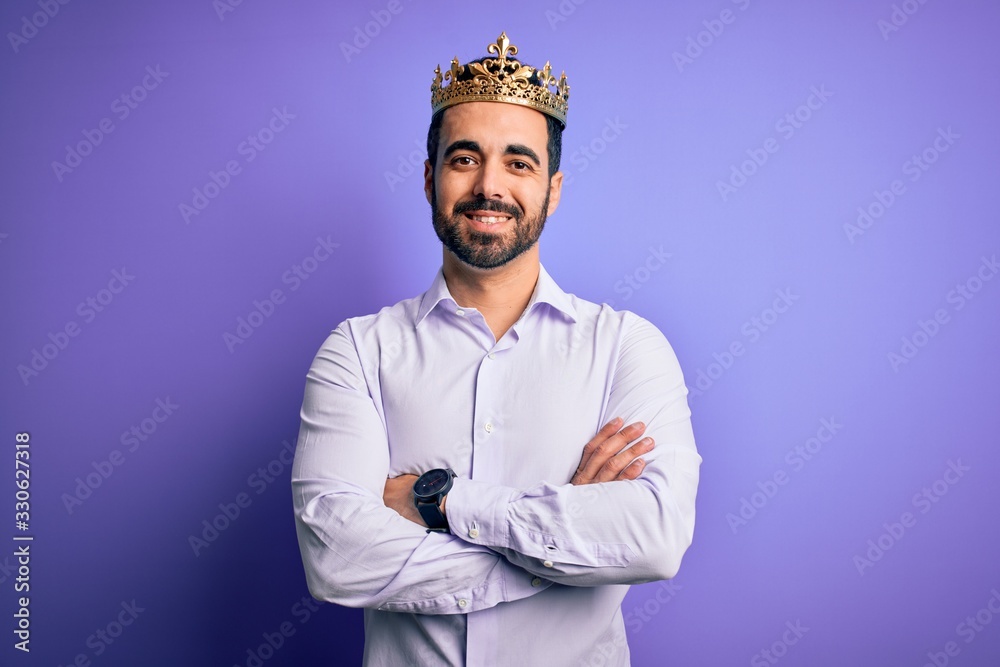 Young handsome man with beard wearing golden crown of king over purple background happy face smiling with crossed arms looking at the camera. Positive person. Young handsome man with beard wearing golden crown of king over purple background happy face smiling with crossed arms looking at the camera. Positive person.