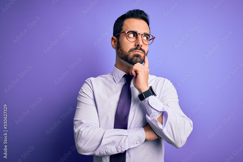 Handsome businessman with beard wearing casual tie and glasses over purple background with hand on chin thinking about question, pensive expression. Smiling with thoughtful face. Doubt concept. Handsome businessman with beard wearing casual tie and glasses over purple background with hand on chin thinking about question, pensive expression. Smiling with thoughtful face. Doubt concept.