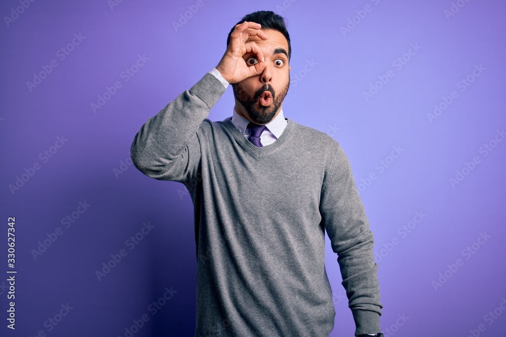 Handsome businessman with beard wearing casual tie standing over purple background doing ok gesture shocked with surprised face, eye looking through fingers. Unbelieving expression.
