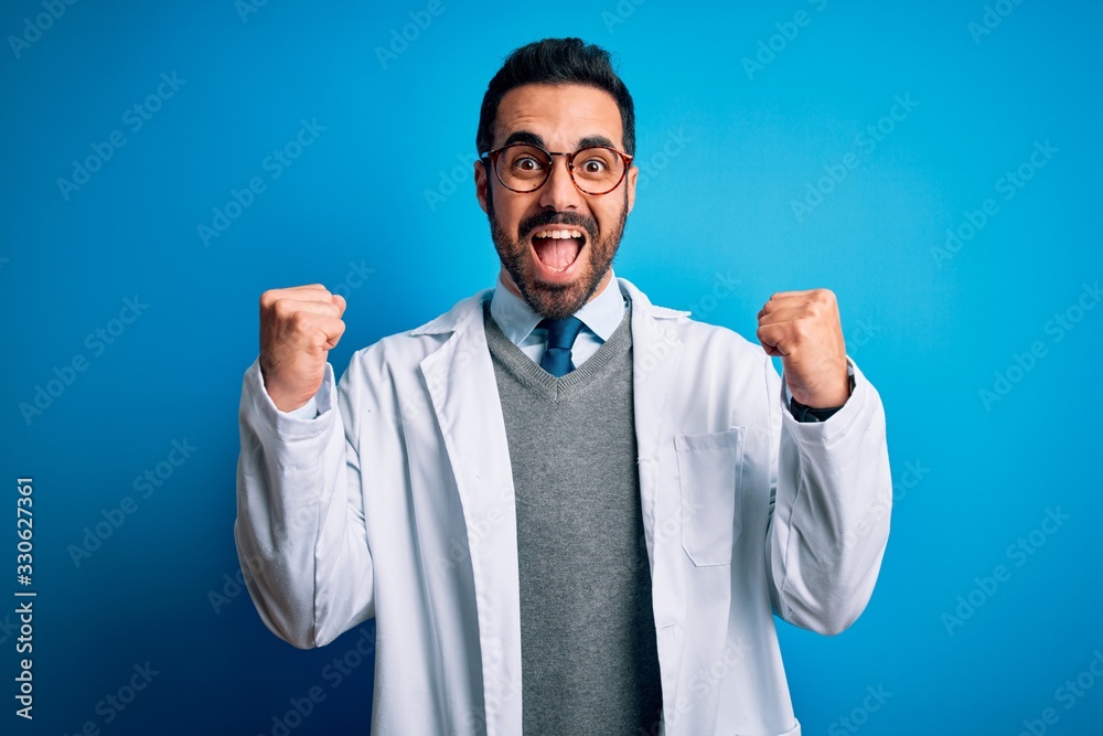 Young handsome doctor man with beard wearing coat and glasses over blue background celebrating surprised and amazed for success with arms raised and open eyes. Winner concept. Young handsome doctor man with beard wearing coat and glasses over blue background celebrating surprised and amazed for success with arms raised and open eyes. Winner concept.
