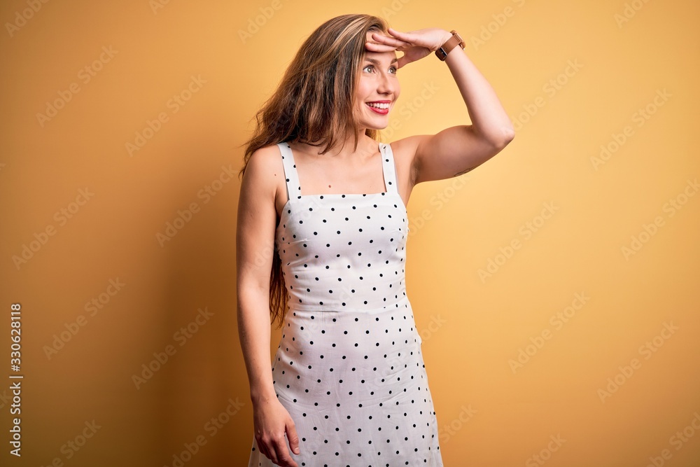 Young beautiful blonde woman on vacation wearing summer dress over yellow background very happy and smiling looking far away with hand over head. Searching concept. Young beautiful blonde woman on vacation wearing summer dress over yellow background very happy and smiling looking far away with hand over head. Searching concept.