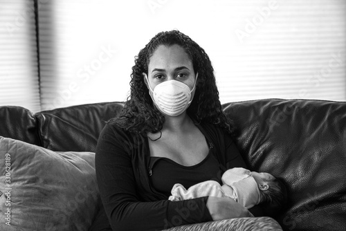 A young African American mother sits on a black leather sofa holding her newborn daughter to her chest wearing a dust mask on her face in hopes of preventing sickness from caronavirus or COVID-19. photo