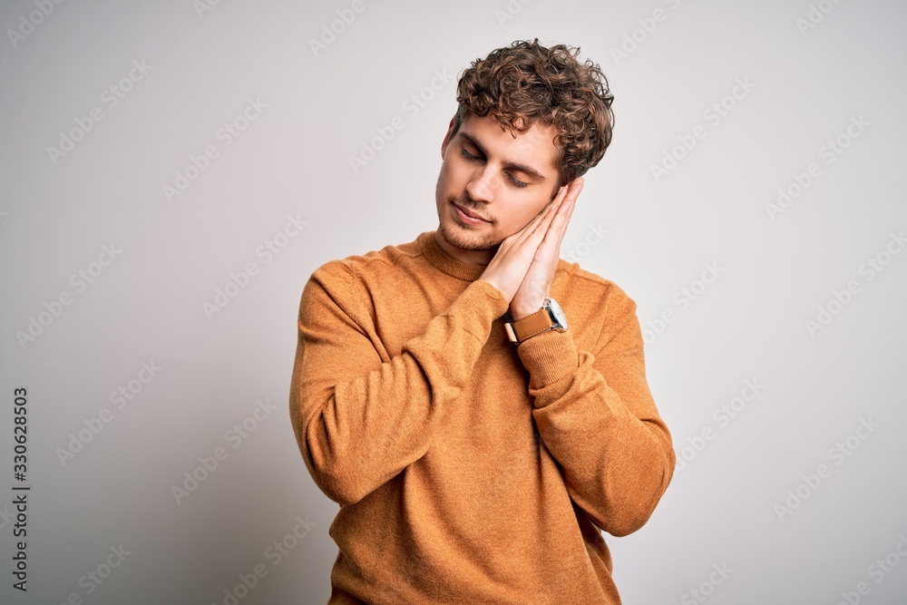 Obraz premium Young blond handsome man with curly hair wearing casual sweater over white background sleeping tired dreaming and posing with hands together while smiling with closed eyes.