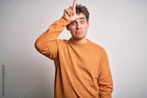 Young blond handsome man with curly hair wearing casual sweater over white background making fun of people with fingers on forehead doing loser gesture mocking and insulting.