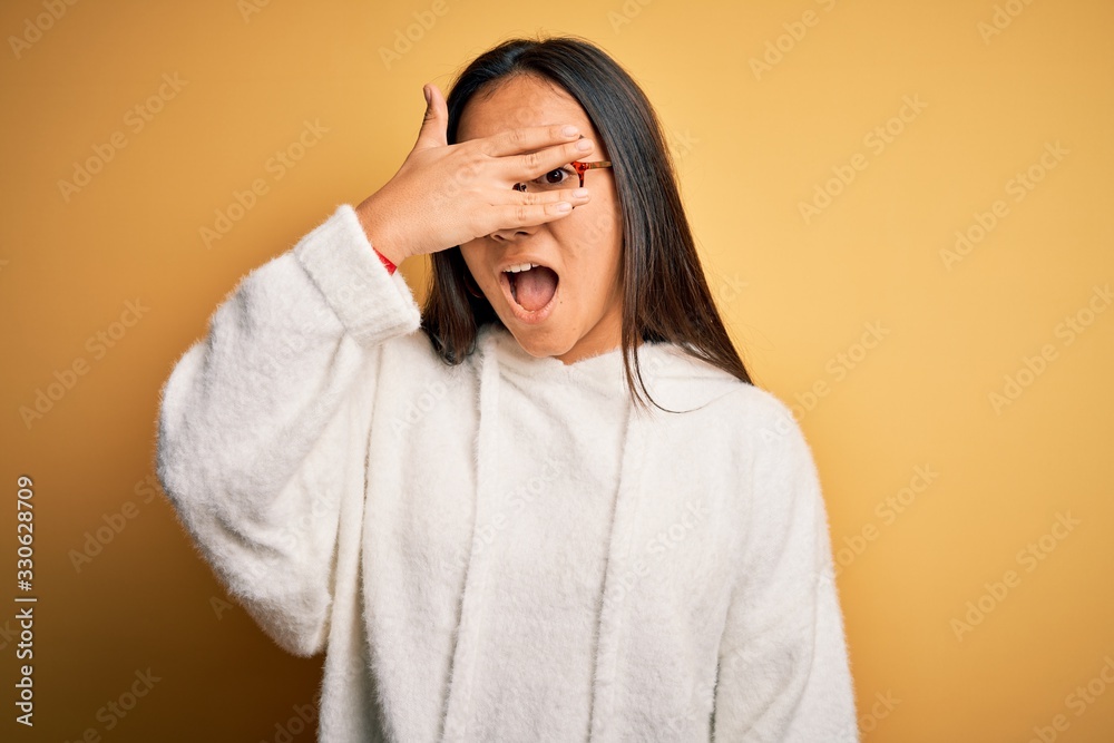 Young beautiful asian woman wearing casual sweater and glasses over yellow background peeking in shock covering face and eyes with hand, looking through fingers with embarrassed expression. Young beautiful asian woman wearing casual sweater and glasses over yellow background peeking in shock covering face and eyes with hand, looking through fingers with embarrassed expression.