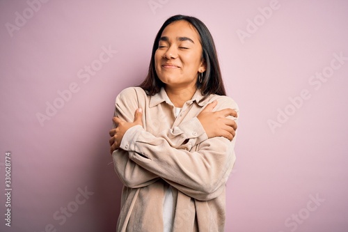 Young beautiful asian woman wearing casual shirt standing over pink background Hugging oneself happy and positive, smiling confident. Self love and self care © Krakenimages.com