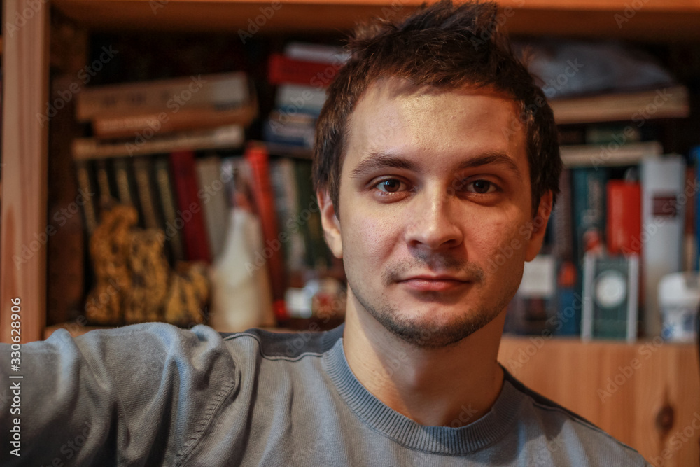 Fototapeta premium Portrait of a young seriously man at home on the background of the bookcase looking to camera.