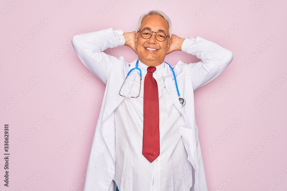 Middle age senior grey-haired doctor man wearing stethoscope and professional medical coat relaxing and stretching, arms and hands behind head and neck smiling happy Middle age senior grey-haired doctor man wearing stethoscope and professional medical coat relaxing and stretching, arms and hands behind head and neck smiling happy