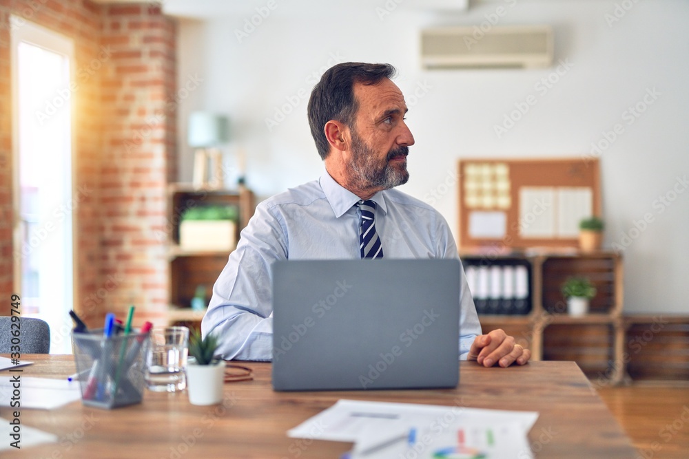 Middle age handsome businessman wearing tie sitting using laptop at the office looking to side, relax profile pose with natural face with confident smile. Middle age handsome businessman wearing tie sitting using laptop at the office looking to side, relax profile pose with natural face with confident smile.