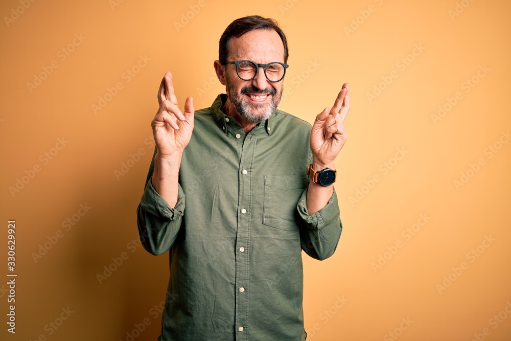 Middle age hoary man wearing casual green shirt and glasses over isolated yellow background gesturing finger crossed smiling with hope and eyes closed. Luck and superstitious concept. Middle age hoary man wearing casual green shirt and glasses over isolated yellow background gesturing finger crossed smiling with hope and eyes closed. Luck and superstitious concept.