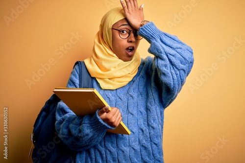 Young African American student woman wearing muslim hijab and backpack holding book surprised with hand on head for mistake, remember error. Forgot, bad memory concept. © Krakenimages.com
