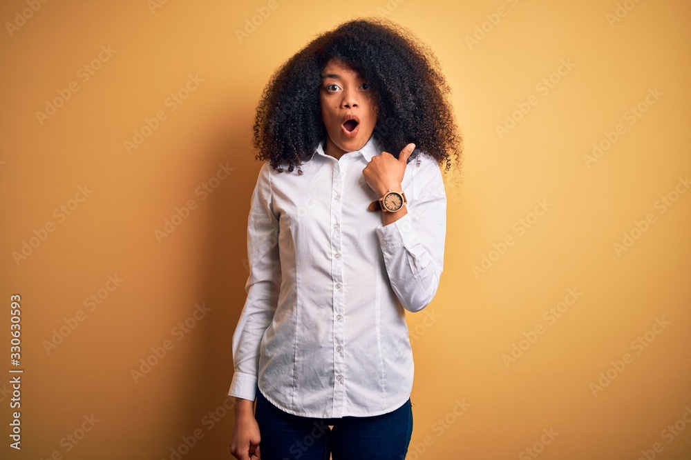 Young beautiful african american elegant woman with afro hair standing over yellow background Surprised pointing with hand finger to the side, open mouth amazed expression. Young beautiful african american elegant woman with afro hair standing over yellow background Surprised pointing with hand finger to the side, open mouth amazed expression.