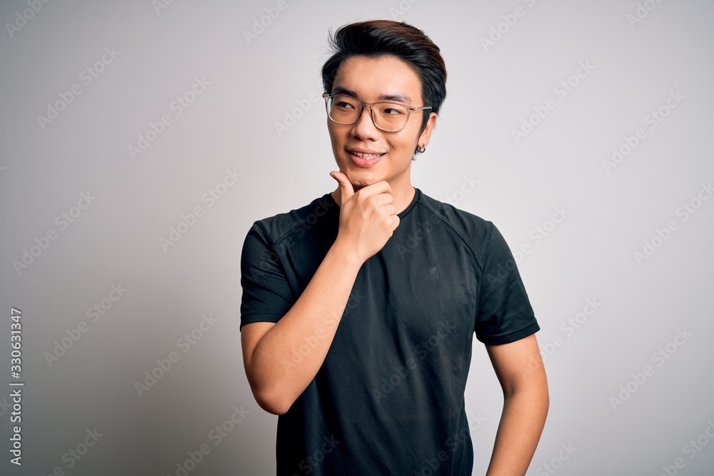 Young handsome chinese man wearing black t-shirt and glasses over white background with hand on chin thinking about question, pensive expression. Smiling with thoughtful face. Doubt concept. Young handsome chinese man wearing black t-shirt and glasses over white background with hand on chin thinking about question, pensive expression. Smiling with thoughtful face. Doubt concept.