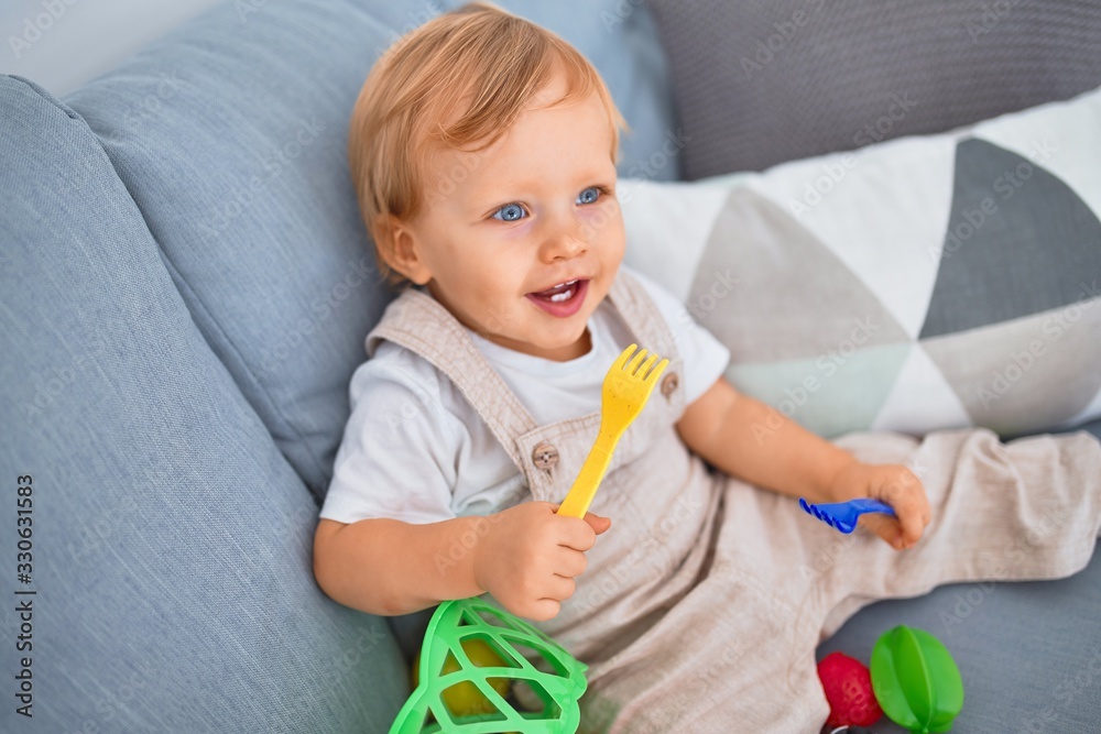 Adorable blonde toddler smiling happy sitting on the sofa playing with plastic meals toys at home Adorable blonde toddler smiling happy sitting on the sofa playing with plastic meals toys at home