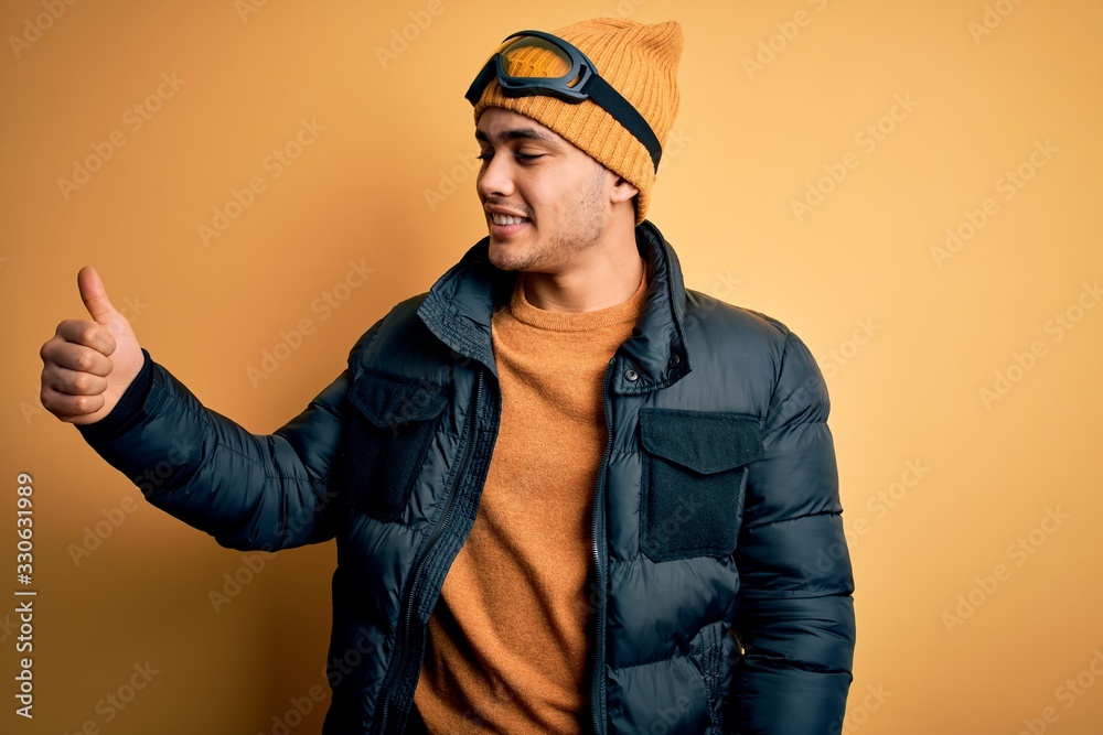 Young brazilian skier man wearing snow sportswear and ski goggles over yellow background Looking proud, smiling doing thumbs up gesture to the side Young brazilian skier man wearing snow sportswear and ski goggles over yellow background Looking proud, smiling doing thumbs up gesture to the side