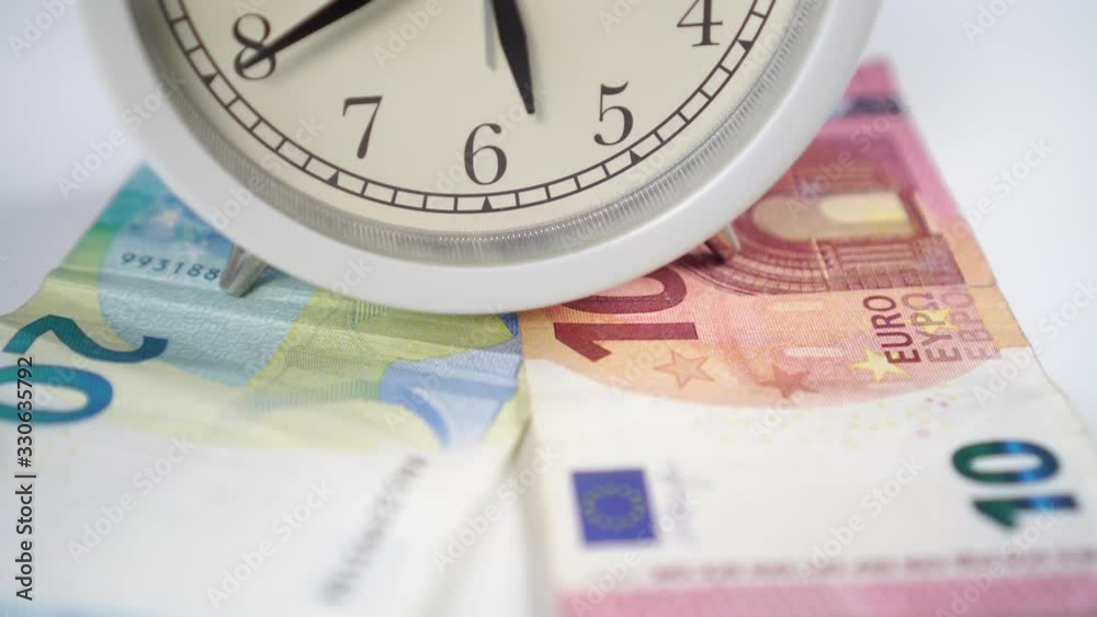 White alarm clock with black arrows and euro banknotes on a white table close-up. Macro shot. Concept of financial savings and loan repayment. White alarm clock with black arrows and euro banknotes on a white table close-up. Macro shot. Concept of financial savings and loan repayment.