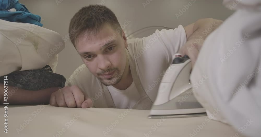 Young confident Caucasian man ironing clothes indoors. Portrait of brunette handsome guy doing housework. Focused on person face. Housekeeping, lifestyle, household. Cinema 4k ProRes HQ. Young confident Caucasian man ironing clothes indoors. Portrait of brunette handsome guy doing housework. Focused on person face. Housekeeping, lifestyle, household. Cinema 4k ProRes HQ.